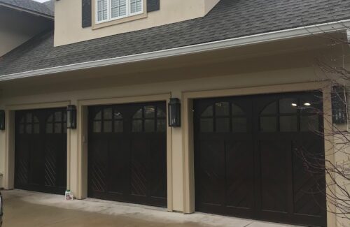 Tan home with a three car garage. Garage doors are painted black and have two windows at the top of each.